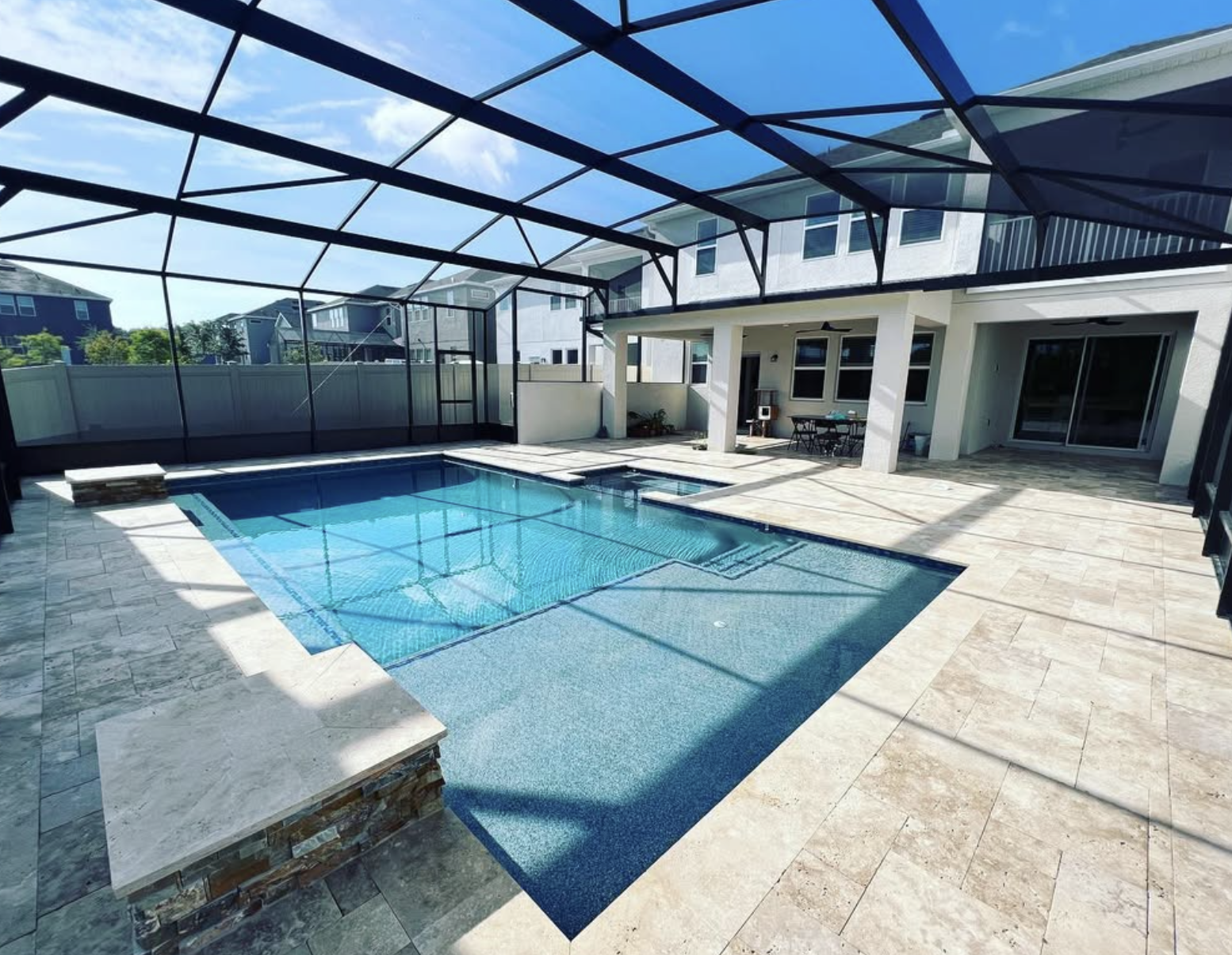 A screened-in backyard pool with stone tile decking, water features, and patio seating next to a two-story house under a blue sky.